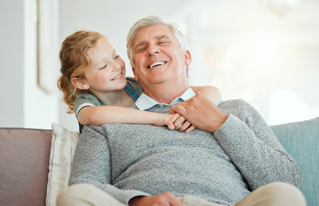 Grandfather and granddaughter laughing together on couch, celebrating Father’s Day joyfully